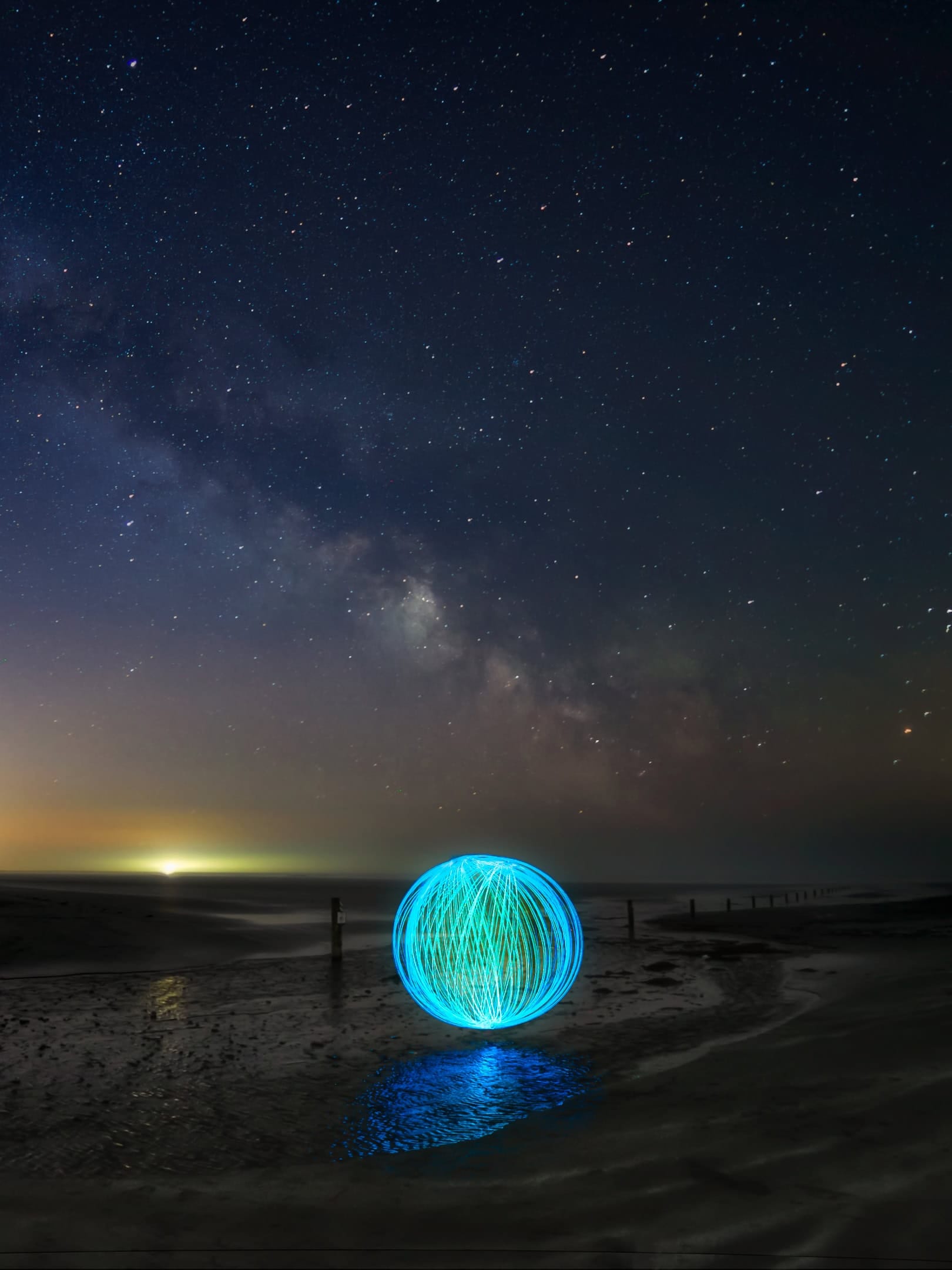 A sphere of light rests on the sandy beach with the Milky Way stretching across the sky