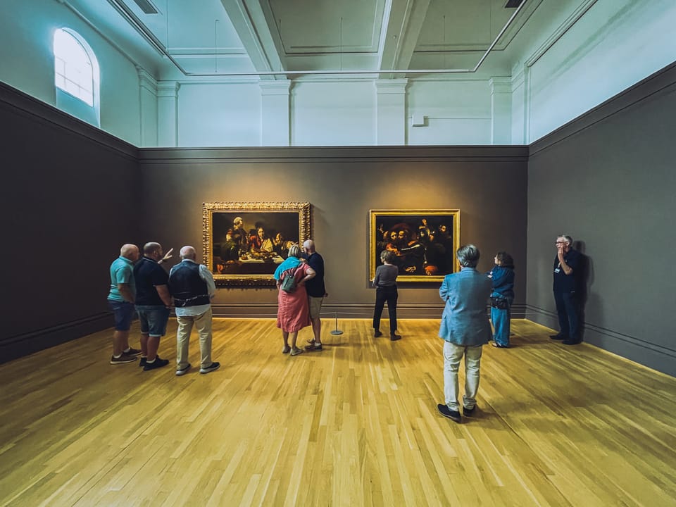 A wide shot of an art exhibit. Two Caravaggio paintings being admired by visitors.