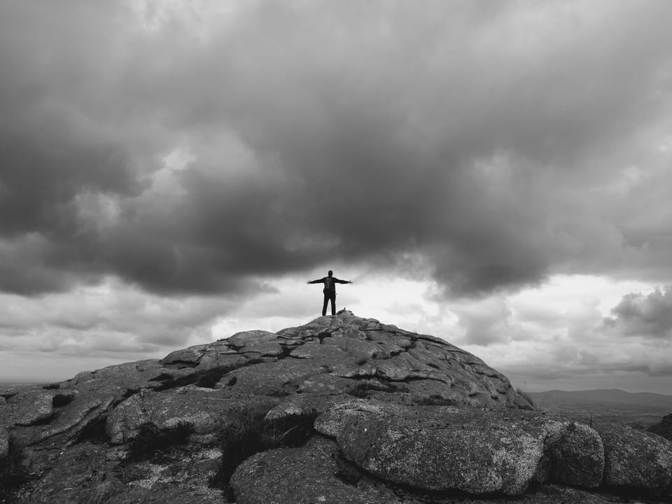 A young man standing at the summit of a mountain, back to the camera with arms stretched out. In black and white.